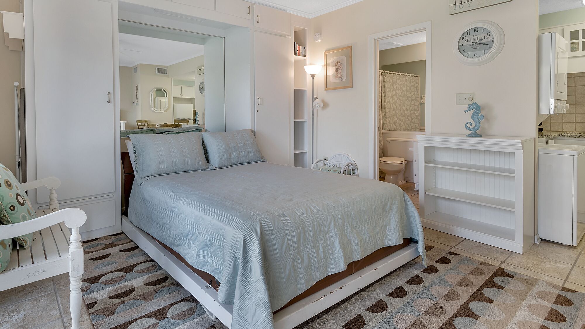 Un dormitorio con cama, una silla, un armario y un baño visible a través de una puerta abierta.