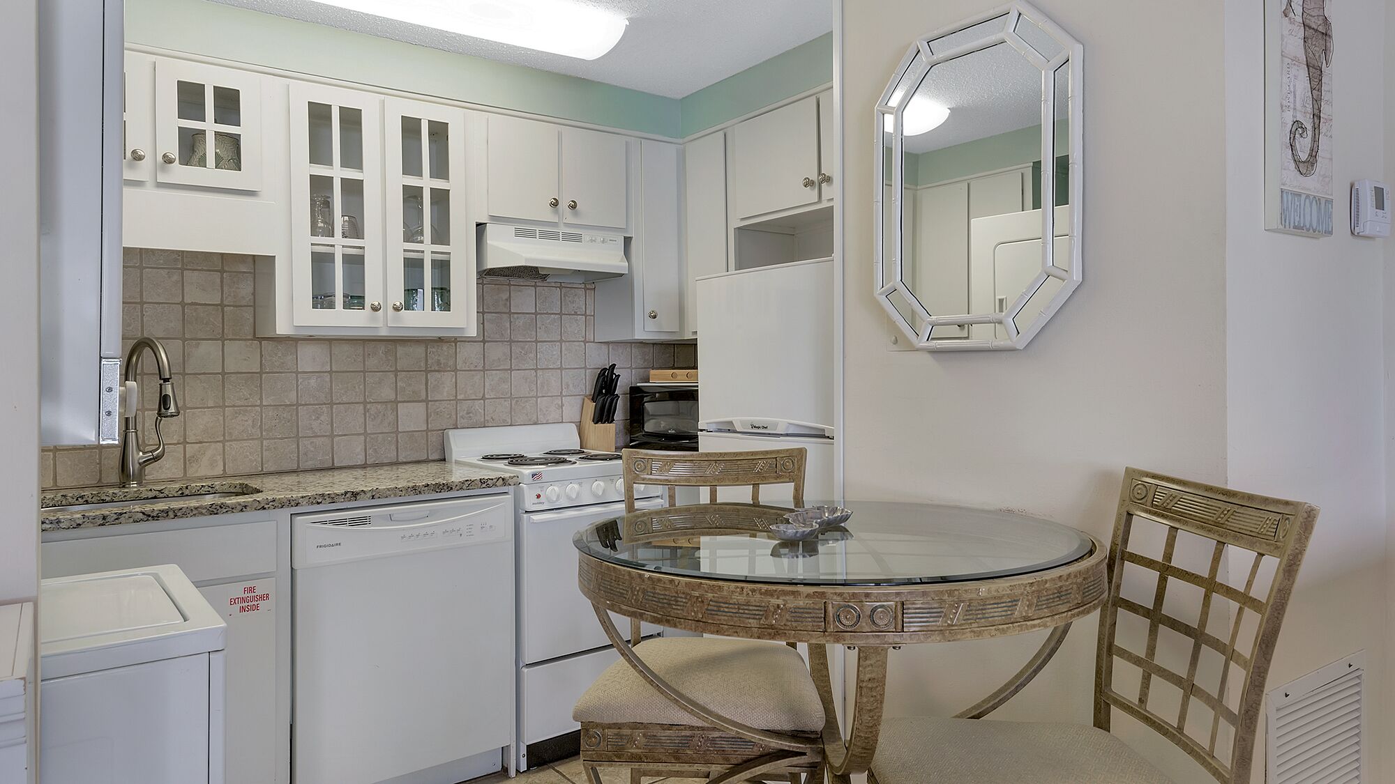 Une cuisine compacte avec des meubles blancs, une cuisinière, un four à micro-ondes et un lave-vaisselle. On y trouve une table ronde avec des chaises et un miroir accroché au mur.