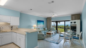 Uma cozinha moderna com armários brancos, uma área de refeições com mesa e cadeiras, um sofá e uma grande janela com vista para o oceano.