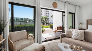 A modern hotel room with a large sliding glass door leading to a balcony, a sofa, a chair, and a small table with decorative items.