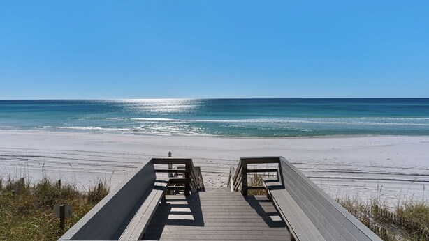 A wooden boardwalk leading to a beach with clear blue water and a sandy shore.