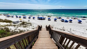 Una scala di legno conduce a una spiaggia con sabbia bianca e ombrelloni blu.