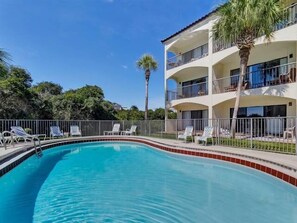 A two-story building with a swimming pool, surrounded by a fence and palm trees.