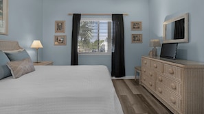 Une chambre à coucher avec un lit, une commode, un téléviseur et une fenêtre avec des rideaux.