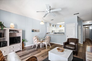 Un salon équipé d’un téléviseur, de meubles blancs, d’un ventilateur de plafond et d’un coin repas avec des chaises.