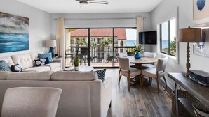Uma sala de estar espaçosa com um sofá grande, uma mesa de jantar com cadeiras, uma televisão e uma varanda com vista para o mar.