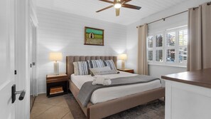 Un dormitorio con cama, dos mesitas de noche, ventilador de techo, ventana con contraventanas y una obra de arte colgada en la pared.