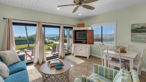 A spacious living room with a large sliding glass door leading to a patio, a ceiling fan, a television, and a dining area with white furniture. - Updated Gulf Front in Seagrove, Direct Sunset View + Free Attraction Tickets! (Santa Rosa Beach)