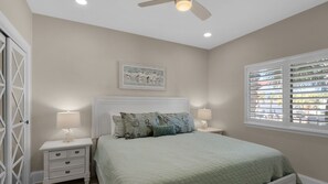 Quarto com cama, duas mesas de cabeceira, ventilador de teto, janela com persianas e um quadro na parede.