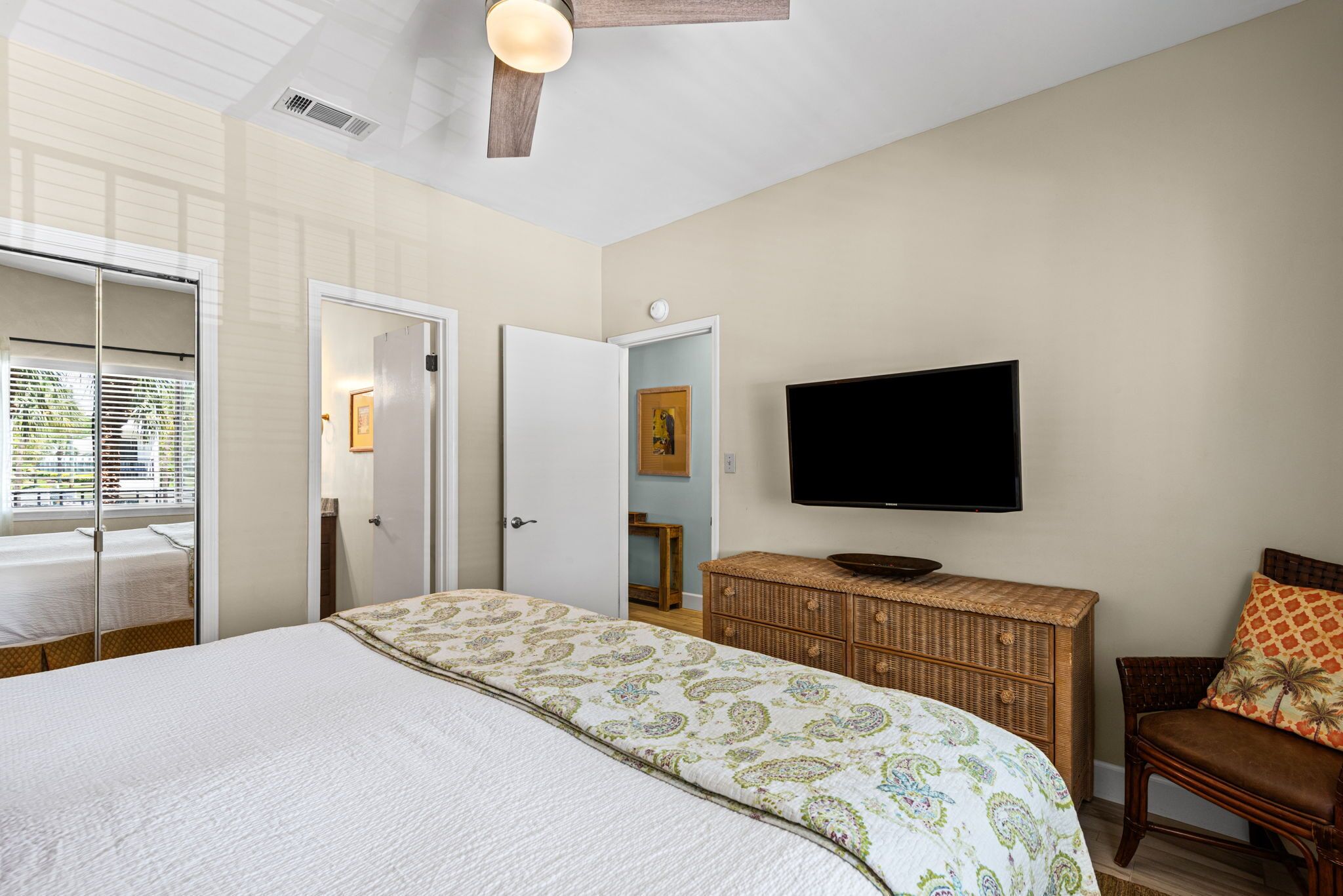 Une chambre à coucher avec un lit, un téléviseur fixé au mur, une commode en osier et une chaise.