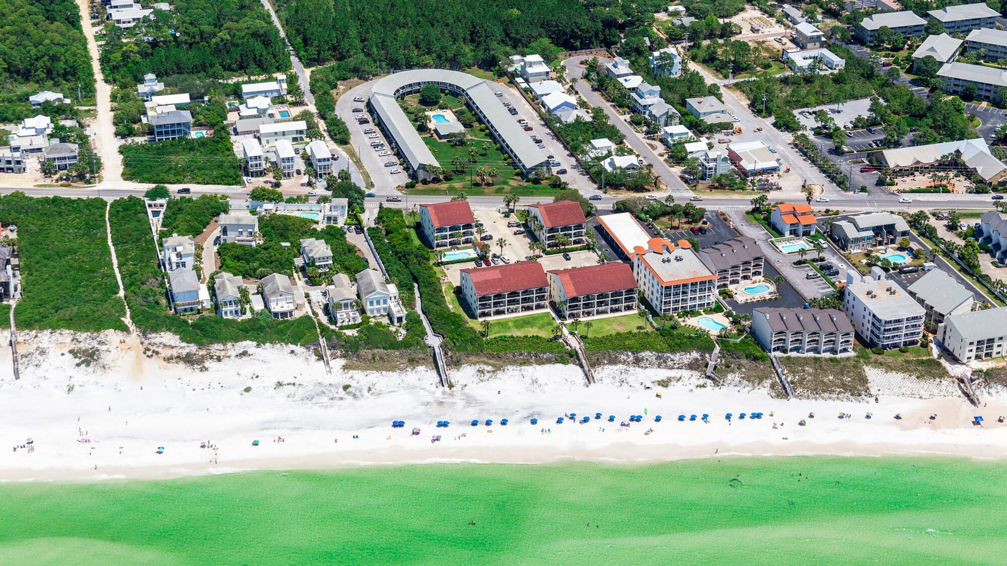 An aerial view of a coastal residential area with beachfront properties, a curved road, and a beach with people.