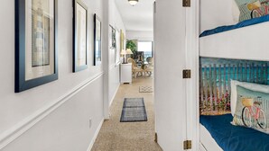 A hallway with framed pictures on the wall, a dining area in the background, and a bed with a blue headboard and a bicycle-themed pillow. - Gulf-Front, Pool in Seagrove, Next Door to Seaside + Free Attraction Tickets! (Santa Rosa Beach)