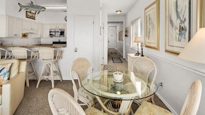 A dining area with wicker chairs and a glass table, a kitchen with white cabinets and a microwave, and a hallway with framed artwork. - Gulf-Front, Pool in Seagrove, Next Door to Seaside + Free Attraction Tickets! (Santa Rosa Beach)