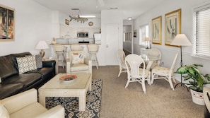 A modern living room with a sofa, coffee table, and chairs, a kitchen area with white cabinets and appliances, and a dining area with a table and chairs.