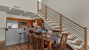 A modern kitchen with wooden cabinets, a stainless steel refrigerator, and a microwave. There is a dining area with a wooden table and chairs, and a staircase with wooden railings.
