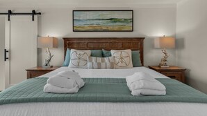 A neatly made bed with a green quilt, two white towels, and decorative pillows. There are two bedside tables with lamps and a painting above the bed.