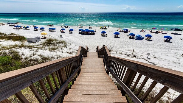 A wooden boardwalk leading to a beach with blue umbrellas and lounge chairs.