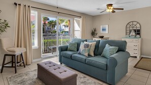 A living room with a teal sofa, a brown ottoman, a white dresser, and a ceiling fan. - Ground Floor, Complex Pool, Near to Beach & Dining + Free Attraction Tickets! (Santa Rosa Beach)