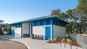 Un bâtiment à la façade bleue et blanche, doté d’un espace extérieur abrité avec des sièges et d’un trottoir bordé de potelets métalliques.