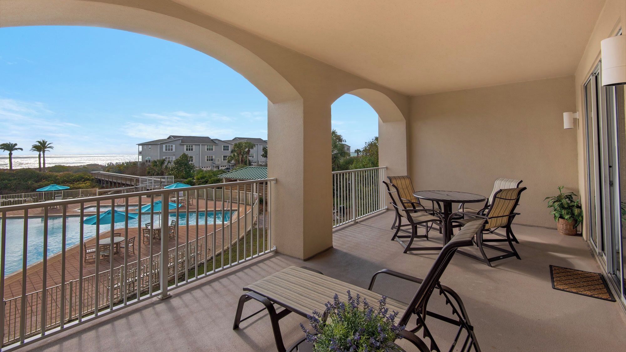 A balcony with a pool, outdoor seating, and a view of a building and beach.