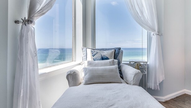 Un dormitorio con vista al océano, una cama con ropa blanca, una silla con cojín y una mesita con un objeto decorativo.