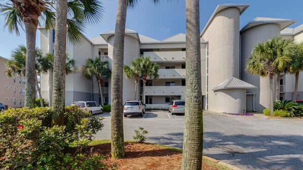 A modern building with a parking lot, palm trees, and a clear sky. - Gulf Front, Pool, Modern Design & Amazing Views + Free Attraction Tickets! (Seacrest)