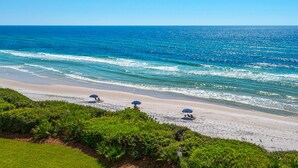A beach with three umbrellas and lounge chairs, a clear view of the ocean, and a grassy area in the foreground. - Gulf Front, Pool, Modern Design & Amazing Views + Free Attraction Tickets! (Seacrest)