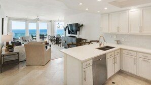 A modern kitchen with white cabinets, a central island with a sink, and a dining area with a view of the ocean. - Gulf Front, Pool, Modern Design & Amazing Views + Free Attraction Tickets! (Seacrest)