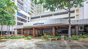 Entrada de hotel com pátio de pedra, área de estar coberta ao ar livre e um edifício de vários andares com varandas.