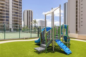 A playground with slides and climbing structures, surrounded by a fence and tall residential buildings.