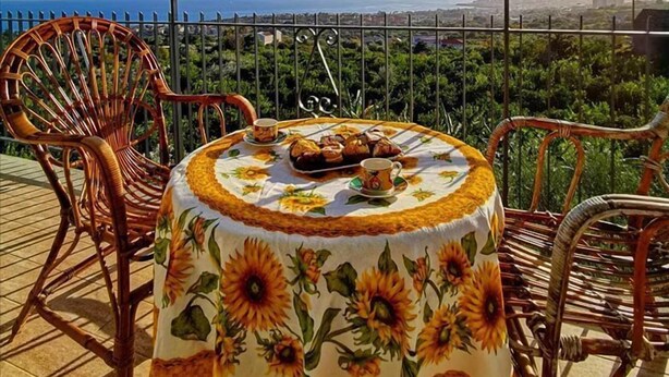 A round table with a sunflower tablecloth, surrounded by wicker chairs, overlooking a cityscape. - Villa Antonio (Aci Castello)