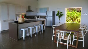Une cuisine moderne avec des meubles blancs, un îlot central doté de tabourets, une table à manger avec des chaises, et une vue sur la verdure par une fenêtre.