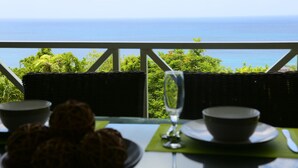 Une table avec un verre, deux bols et une assiette de biscuits, donnant sur un paysage verdoyant et luxuriant et sur la mer.