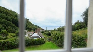A view through a window of a rural landscape with a house, lush greenery, and a distant body of water. - NEW 2021, Idyllic Cottage in Polkerris,  by the beach, sea views, near Fowey. (Par)