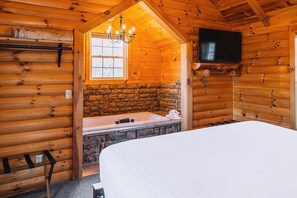 Ein Blockhauszimmer mit Kamin aus Stein, einem Bett, einem an der Wand befestigten Fernseher und einer Kristallleuchtre.