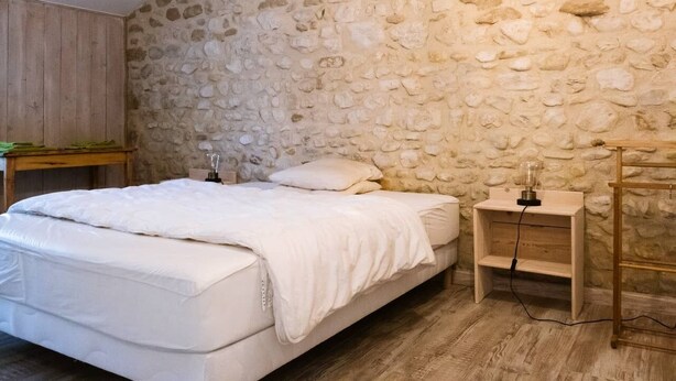 A bedroom with a stone wall, a wooden bed, a bedside table with a lamp, and a wooden nightstand. - abundance, Stone house with land of 2000m2 at the foot of the vercors (La Baume-Cornillane)