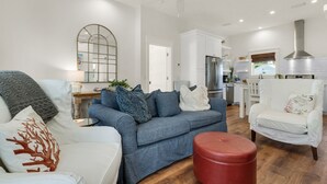 Ein modernes Wohnzimmer mit einem grauen Sofa, einem roten Hocker und einem weißen Sessel. Im Hintergrund ist eine Küche mit weißen Schränken und Edelstahlgeräten zu sehen.
