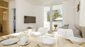 A modern dining area with a wooden table set for four, a flat-screen TV mounted on the wall, and a balcony with a view of trees and a building. - Quartier39 (Sankt Johann in Tirol)