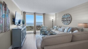 A spacious living room with a balcony, a large wall clock, and a view of the sea.