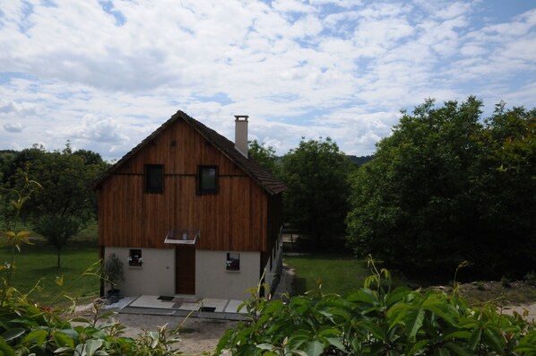 A wooden house with a sloped roof and a chimney, surrounded by greenery.