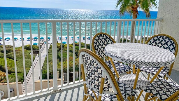 A balcony with a view of the beach and ocean. There are two wicker chairs and a small table.