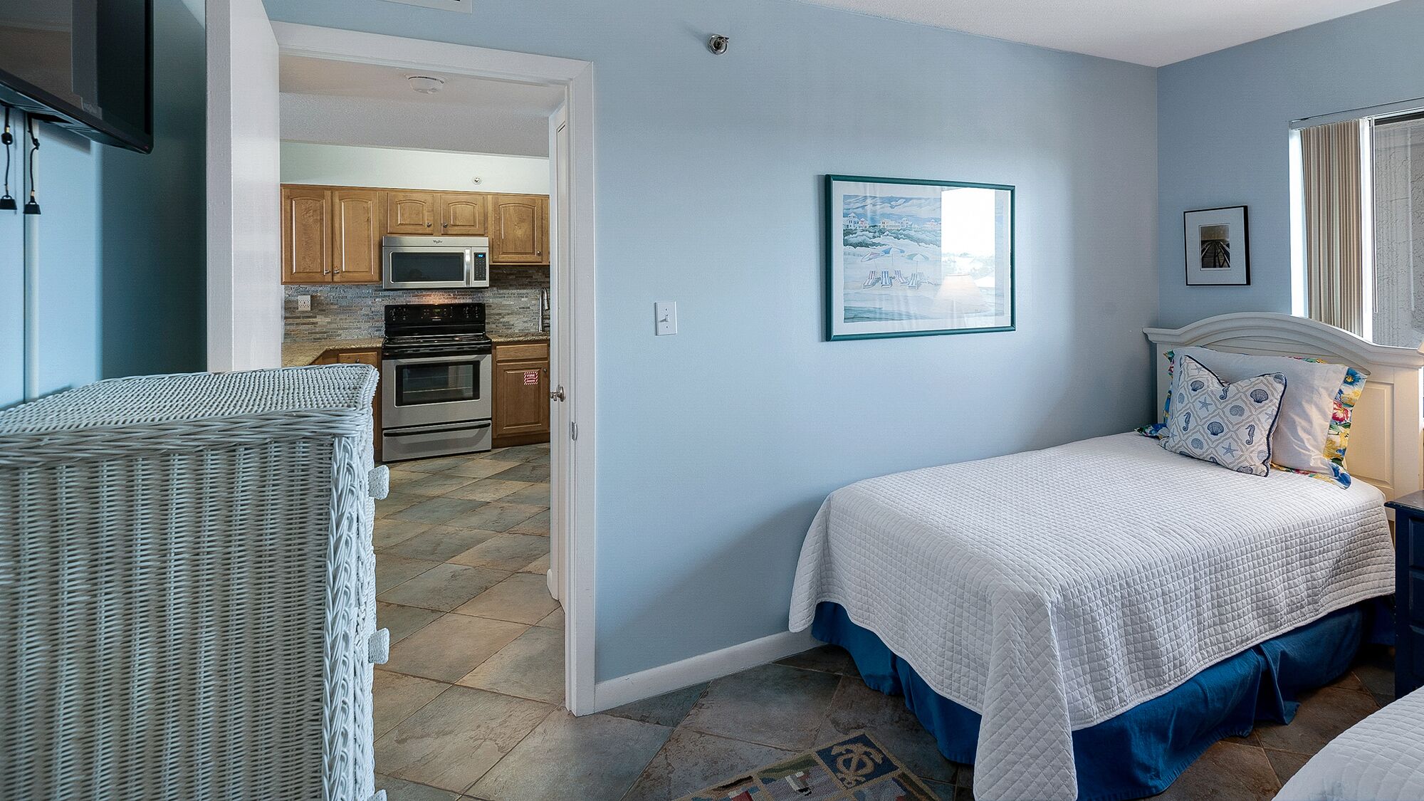 Une chambre avec un lit, une chaise en osier, une cuisine à l’arrière-plan et un tableau encadré au mur.