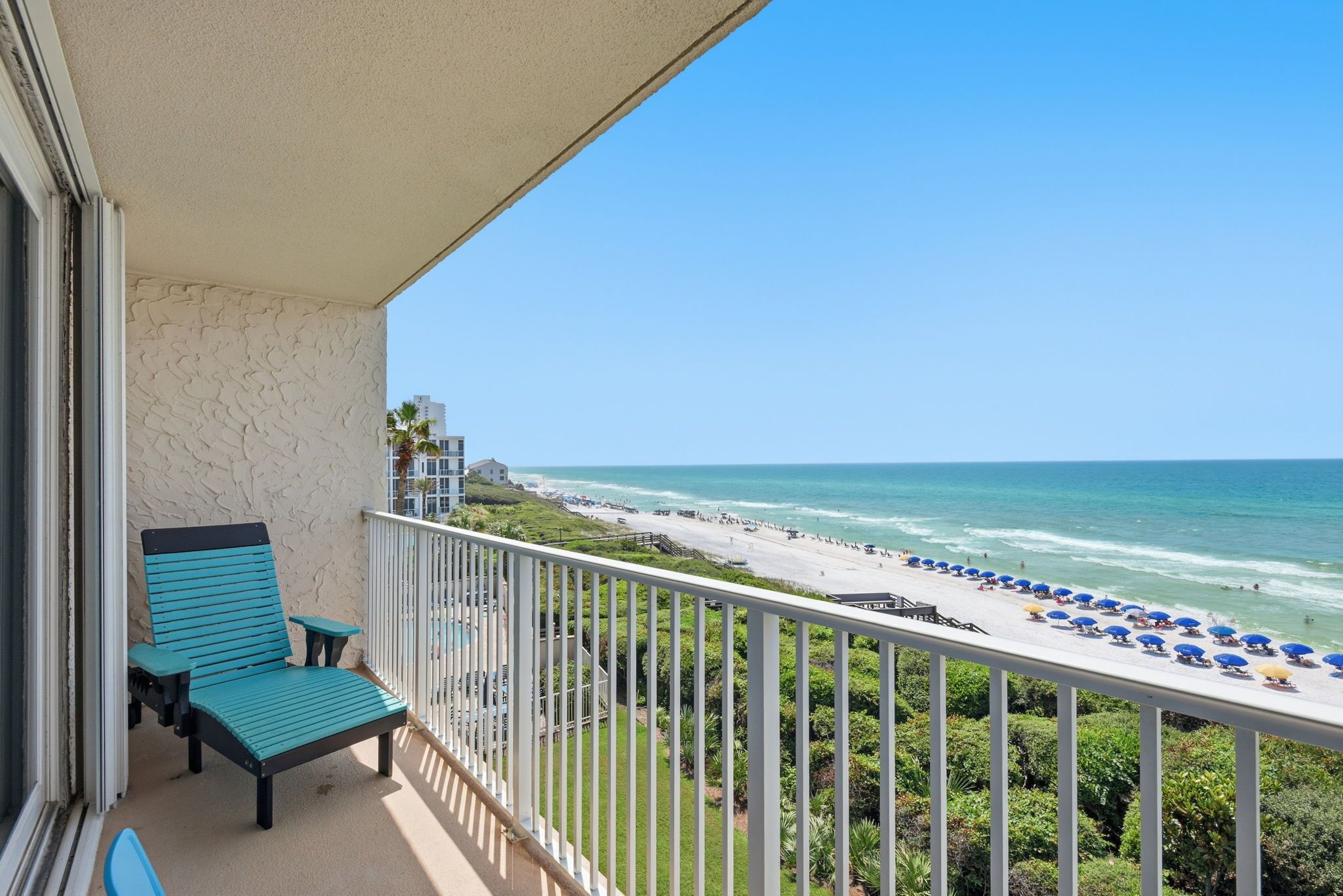 A balcony with a view of a beach, a pool, and a clear sky.