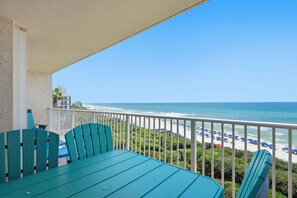 A balcony with a view of the beach, a table, chairs, and a railing.
