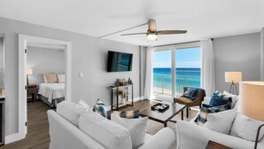 Ein modernes Wohnzimmer mit großem Fenster zum Meer, Deckenventilator und einem an der Wand montierten Flachbildfernseher.