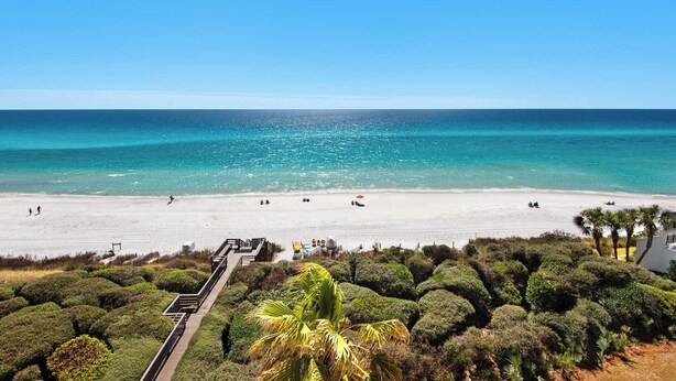 Ein Strand mit einer Holzpromenade, die zum Ufer führt, umgeben von dichter Strauchvegetation.