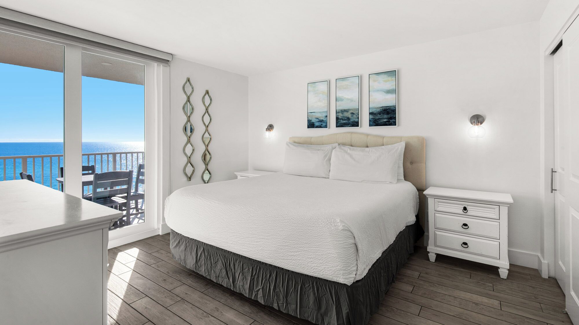 A bedroom with a large bed, a nightstand, a dresser, and a view of the ocean.