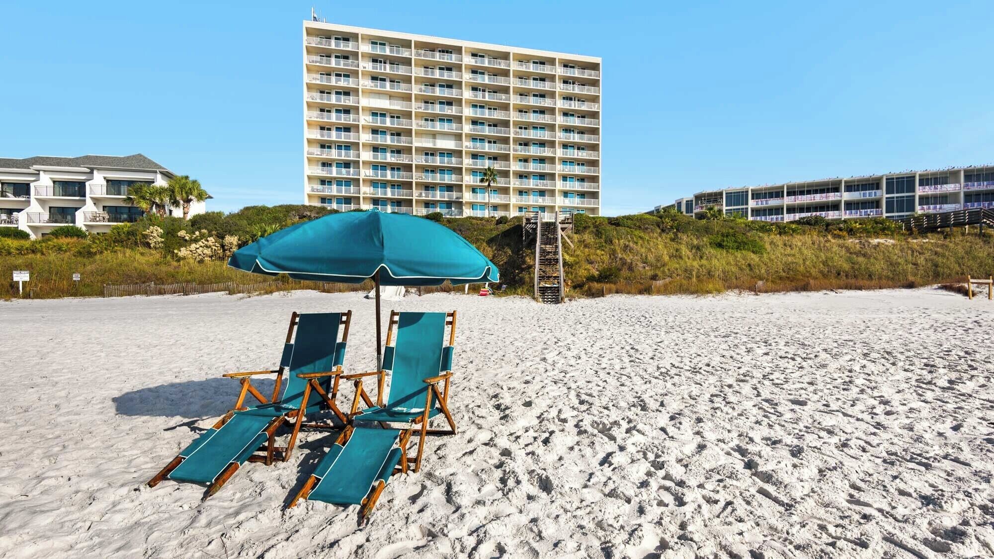 Una spiaggia con due file di sedie a sdraio e un ombrellone grande, un edificio alto sullo sfondo e una scala che conduce verso l'edificio.