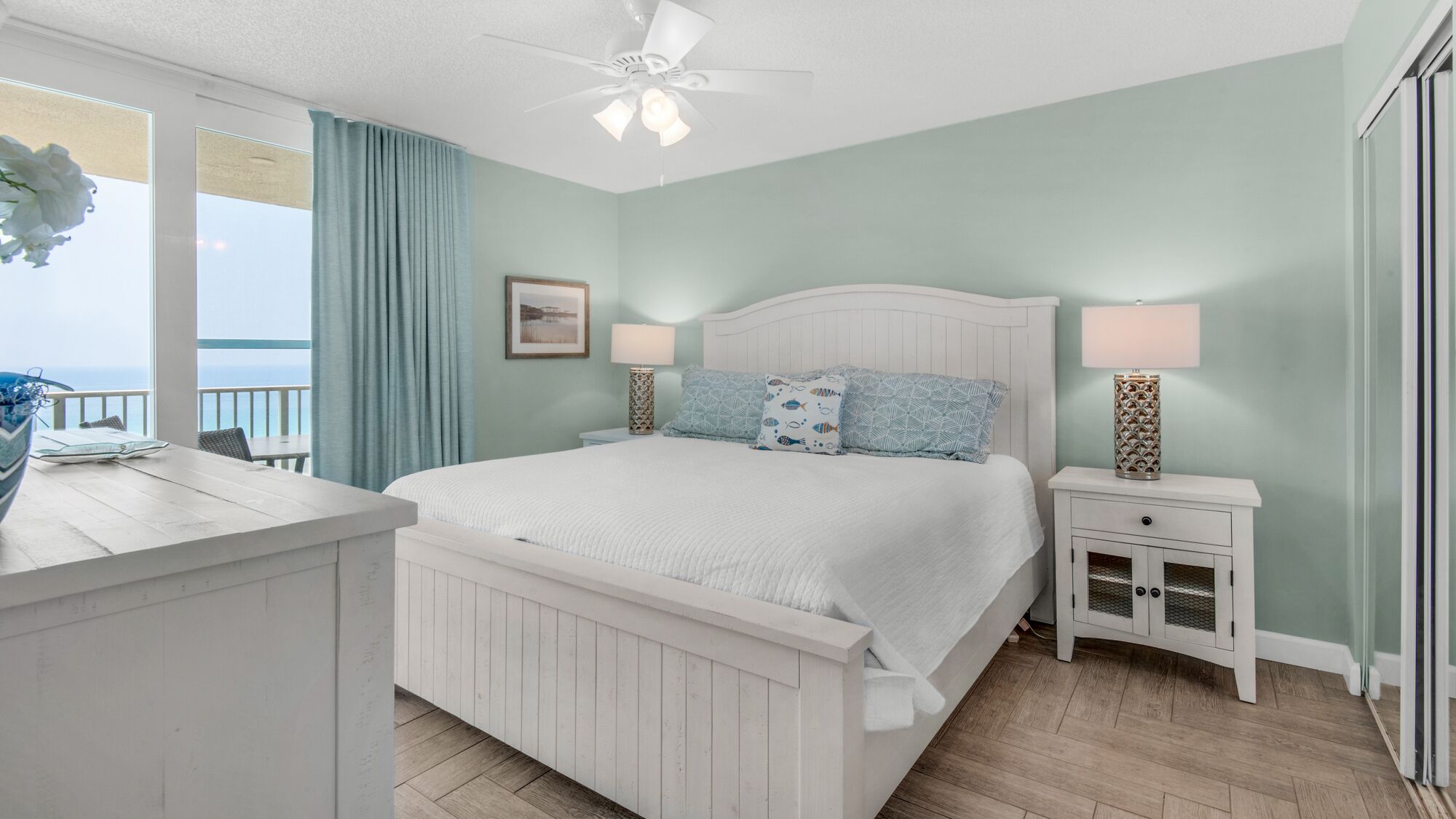 A bedroom with a white bed, two nightstands, a ceiling fan, and a view of the ocean.