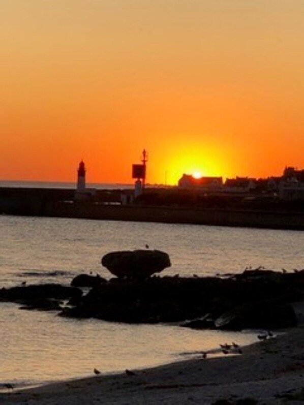 Un coucher de soleil sur une zone côtière, avec un phare et une jetée qui s’avance dans l’eau.